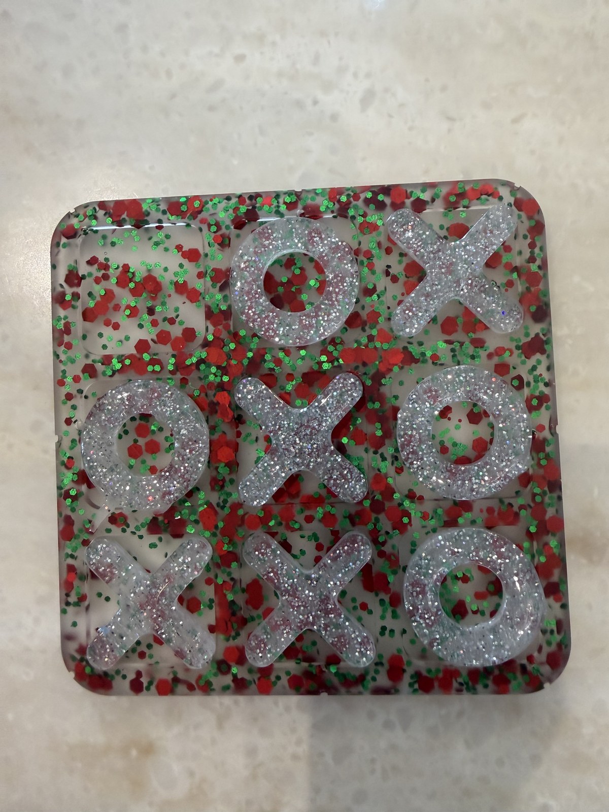 Square resin tic-tac-toe board with red and green glitter and silver glitter X and O pieces on a countertop